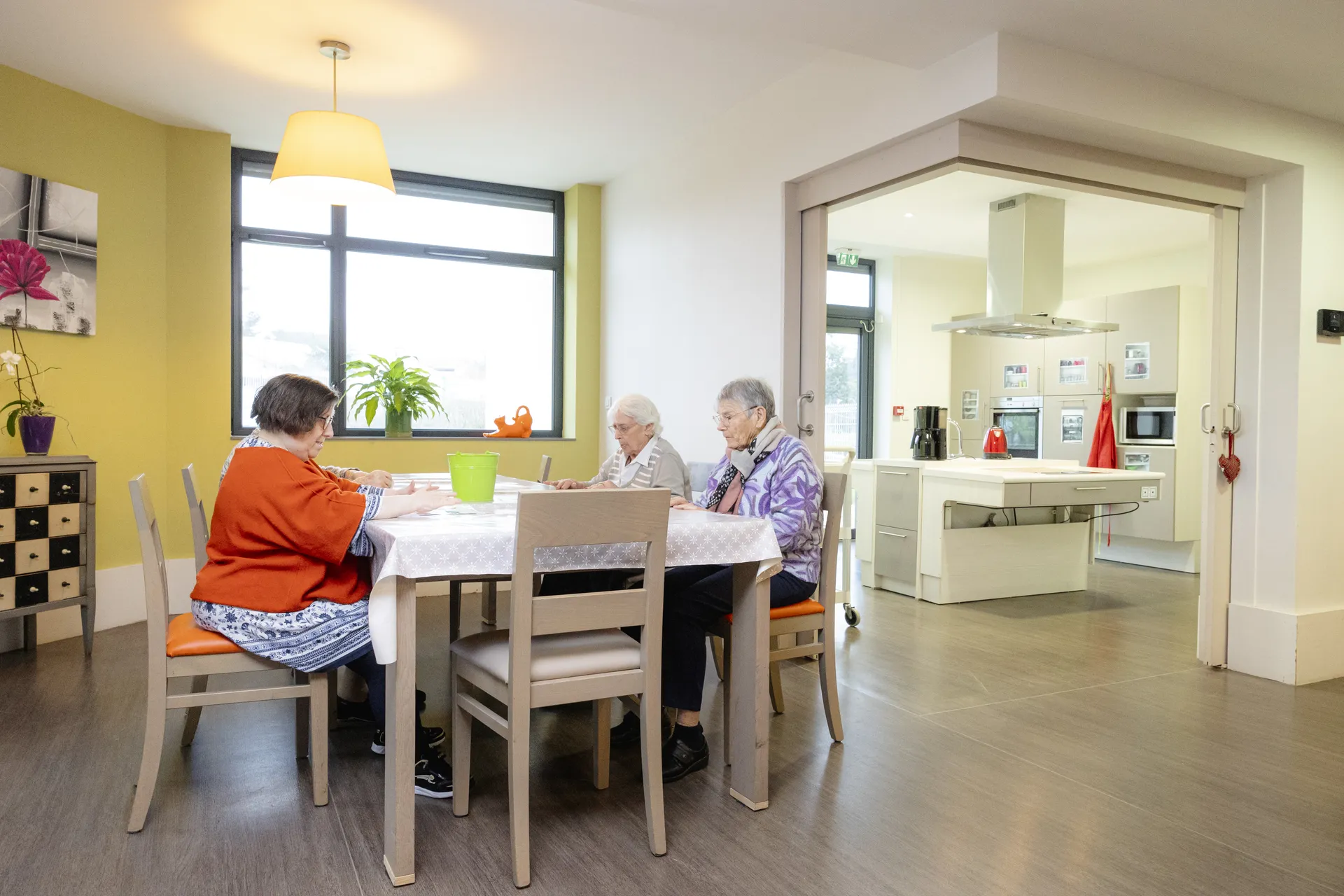 Espace de vie avec coin repas et cuisine ouverte où résidents et accompagnatrice partagent un moment convivial à l'EHPAD Le Clos du Moulin Divatte-sur-Loire